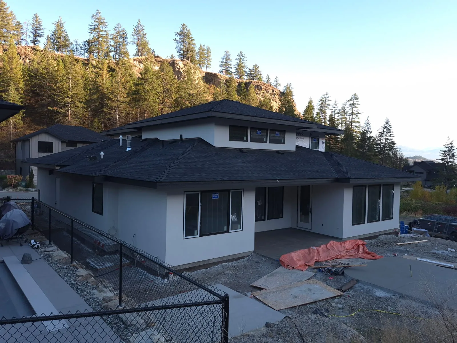 Elevated view of the finished Lot 42 home and rear patio from behind a chain-link fence, pine hillside beyond.